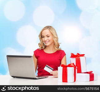 christmas, holidays, technology and shopping concept - smiling woman in red blank shirt with gift boxes, credit card and laptop computer over blue lights background