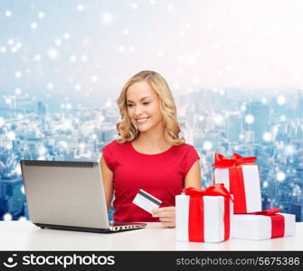 christmas, holidays, technology and shopping concept - smiling woman in red blank shirt with gift boxes, credit card and laptop computer over blue lights background