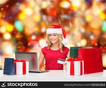 christmas, holidays, technology and shopping concept - smiling woman in santa helper hat with gifts, credit card and laptop computer over red lights background