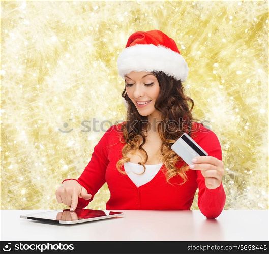christmas, holidays, technology and shopping concept - smiling woman in santa helper hat with credit card and tablet pc computer over yellow lights background