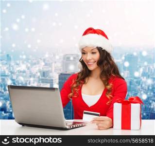 christmas, holidays, technology and shopping concept - smiling woman in santa helper hat with gift box, credit card and laptop computer over snowy city background