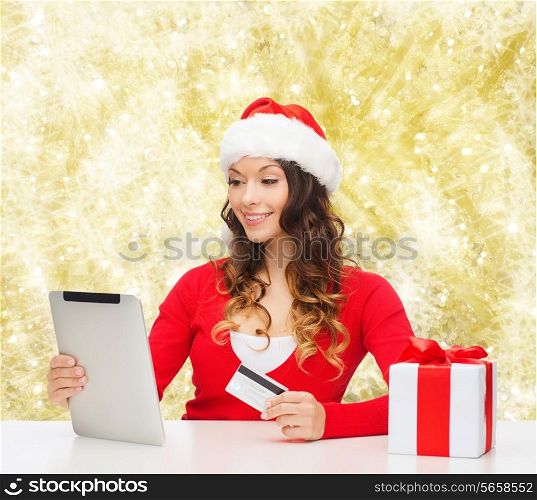 christmas, holidays, technology and shopping concept - smiling woman in santa helper hat with gift box, credit card and tablet pc computer over yellow lights background
