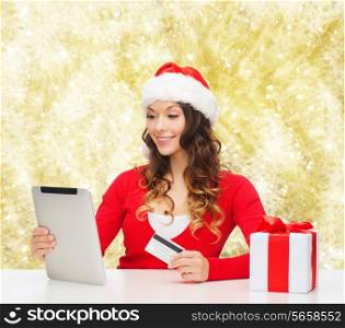 christmas, holidays, technology and shopping concept - smiling woman in santa helper hat with gift box, credit card and tablet pc computer over yellow lights background