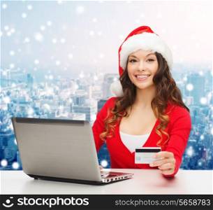christmas, holidays, technology and shopping concept - smiling woman in santa helper hat with credit card and laptop computer over snowy city background