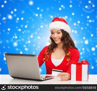 christmas, holidays, technology and shopping concept - smiling woman in santa helper hat with gift box, credit card and laptop computer over blue snowing background
