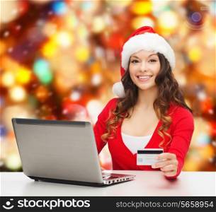 christmas, holidays, technology and shopping concept - smiling woman in santa helper hat with credit card and laptop computer over red lights background