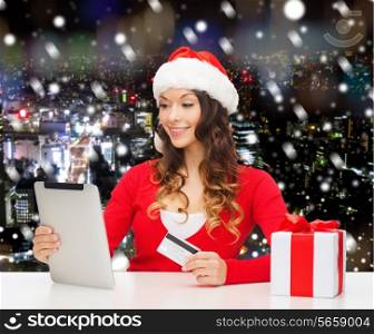 christmas, holidays, technology and shopping concept - smiling woman in santa helper hat with gift box, credit card and tablet pc computer over snowy night city background