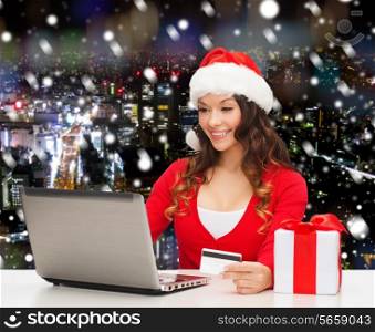christmas, holidays, technology and shopping concept - smiling woman in santa helper hat with gift box, credit card and laptop computer over snowy night city background