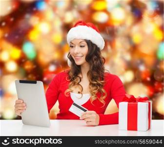 christmas, holidays, technology and shopping concept - smiling woman in santa helper hat with gift box, credit card and tablet pc computer over red lights background