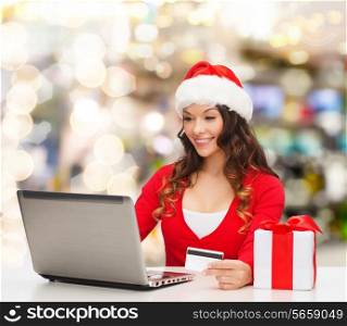 christmas, holidays, technology and shopping concept - smiling woman in santa helper hat with gift box, credit card and laptop computer over lights background