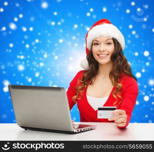 christmas, holidays, technology and shopping concept - smiling woman in santa helper hat with credit card and laptop computer over blue snowing background