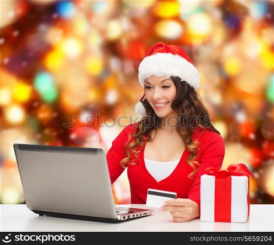 christmas, holidays, technology and shopping concept - smiling woman in santa helper hat with gift box, credit card and laptop computer over red lights background