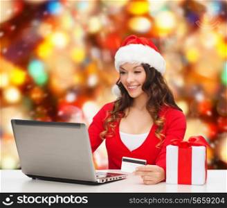 christmas, holidays, technology and shopping concept - smiling woman in santa helper hat with gift box, credit card and laptop computer over red lights background