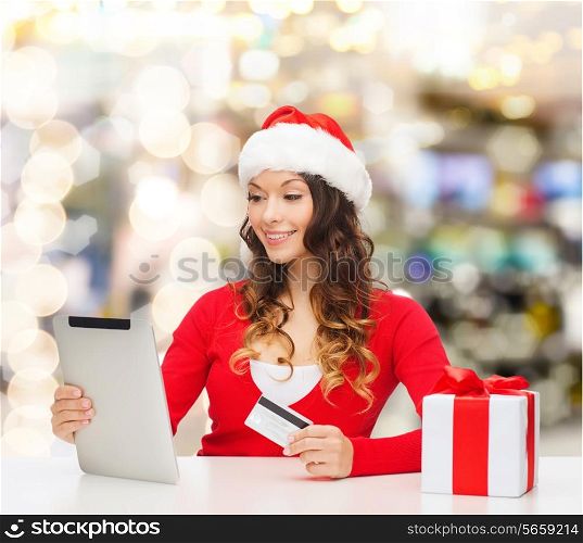 christmas, holidays, technology and shopping concept - smiling woman in santa helper hat with gift box, credit card and tablet pc computer over lights background