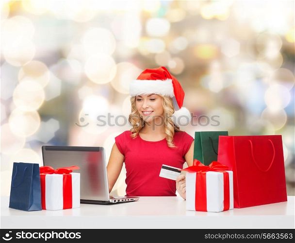 christmas, holidays, technology and shopping concept - smiling woman in santa helper hat with gifts, credit card and laptop computer over lights background