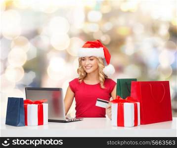 christmas, holidays, technology and shopping concept - smiling woman in santa helper hat with gifts, credit card and laptop computer over lights background