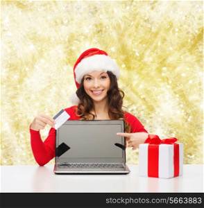 christmas, holidays, technology and shopping concept - smiling woman in santa helper hat with gift box, credit card and laptop computer pointing finger over yellow lights background
