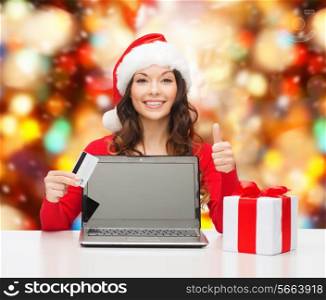 christmas, holidays, technology and shopping concept - smiling woman in santa helper hat with credit card, gift box and laptop computer showing thumbs up gesture over red lights background