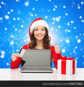 christmas, holidays, technology and shopping concept - smiling woman in santa helper hat with credit card, gift box and laptop computer showing thumbs up gesture over blue snowing background
