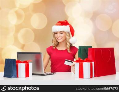 christmas, holidays, technology and shopping concept - smiling woman in santa helper hat with gifts, credit card and laptop computer over beige lights background