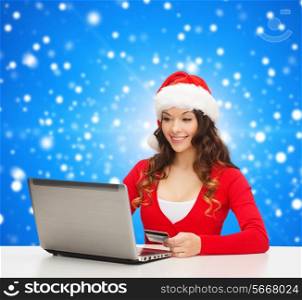 christmas, holidays, technology and shopping concept - smiling woman in santa helper hat with credit card and laptop computer over blue snowing background