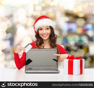 christmas, holidays, technology and shopping concept - smiling woman in santa helper hat with gift box, credit card and laptop computer pointing finger over lights background