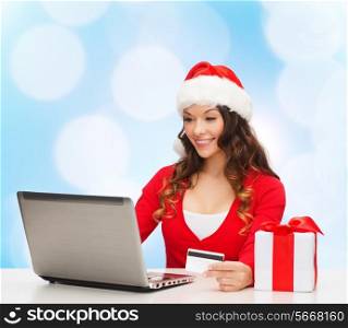 christmas, holidays, technology and shopping concept - smiling woman in santa helper hat with gift box, credit card and laptop computer over blue lights background