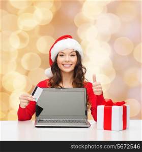 christmas, holidays, technology and shopping concept - smiling woman in santa helper hat with credit card, gift box and laptop computer showing thumbs up gesture over beige lights background