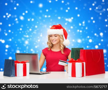 christmas, holidays, technology and shopping concept - smiling woman in santa helper hat with gifts, credit card and laptop computer over blue snowy background