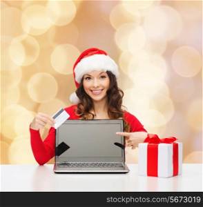 christmas, holidays, technology and shopping concept - smiling woman in santa helper hat with gift box, credit card and laptop computer pointing finger over beige lights background