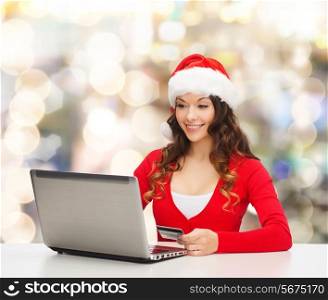 christmas, holidays, technology and shopping concept - smiling woman in santa helper hat with credit card and laptop computer over lights background