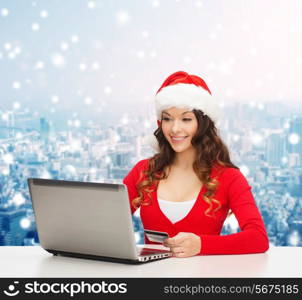 christmas, holidays, technology and shopping concept - smiling woman in santa helper hat with credit card and laptop computer over snowy city background