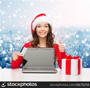 christmas, holidays, technology and shopping concept - smiling woman in santa helper hat with credit card, gift box and laptop computer showing thumbs up gesture over snowy city background