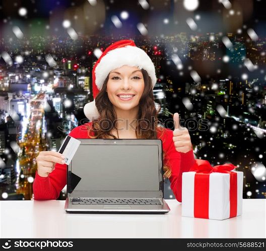 christmas, holidays, technology and shopping concept - smiling woman in santa helper hat with credit card, gift box and laptop computer showing thumbs up gesture over snowy night city background