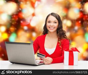 christmas, holidays, technology and shopping concept - smiling woman with credit card, gift box and laptop computer over red lights background