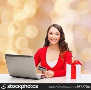 christmas, holidays, technology and shopping concept - smiling woman with credit card, gift box and laptop computer over beige lights background