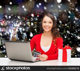 christmas, holidays, technology and shopping concept - smiling woman with credit card, gift box and laptop computer over snowy night city background