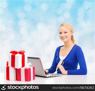 christmas, holidays, technology and shopping concept - smiling woman with gift boxes, credit card and laptop computer over blue lights background