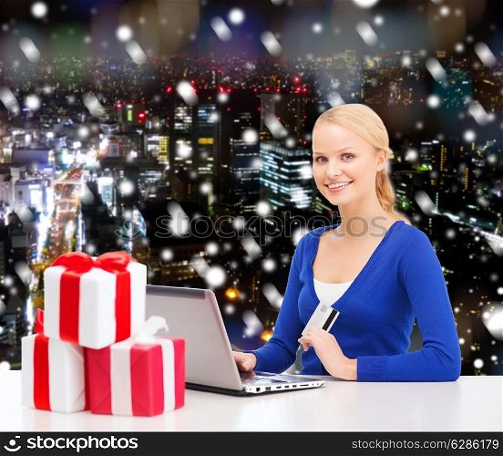 christmas, holidays, technology and shopping concept - smiling woman with gift boxes, credit card and laptop computer over snowy night city background