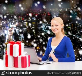 christmas, holidays, technology and shopping concept - smiling woman with gift boxes, credit card and laptop computer over snowy night city background