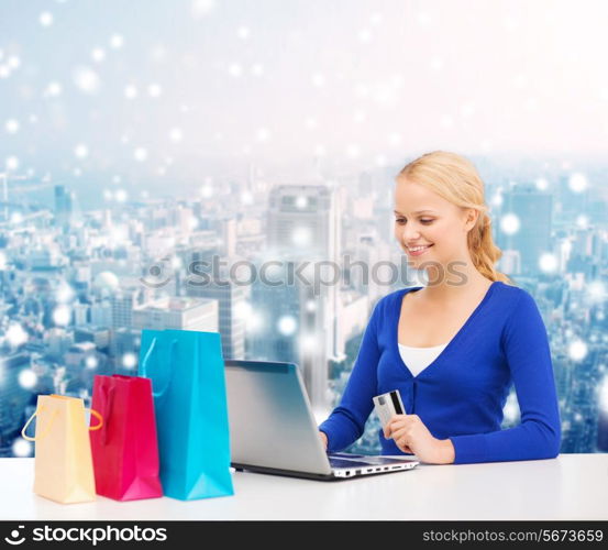 christmas, holidays, technology and shopping concept - smiling woman with shopping bags, credit card and laptop computer over snowy city background