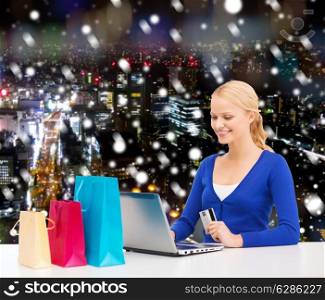 christmas, holidays, technology and shopping concept - smiling woman with shopping bags, credit card and laptop computer over snowy night city background