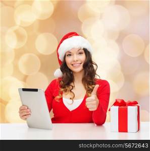 christmas, holidays, technology, gesture and people concept - smiling woman in santa helper hat with gift box and tablet pc computer showing thumbs up over beige lights background