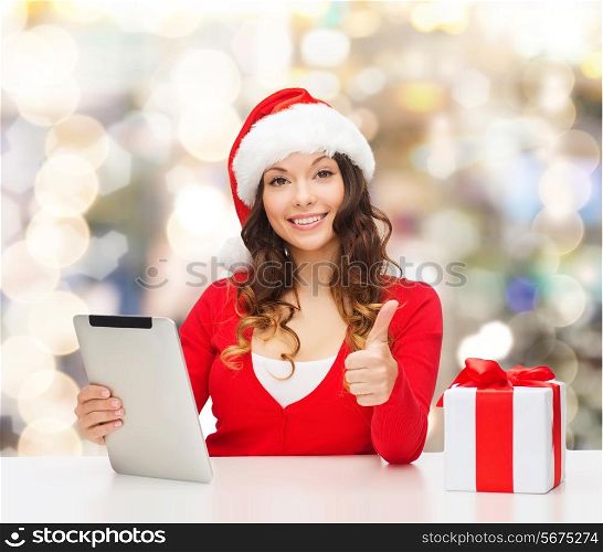 christmas, holidays, technology, gesture and people concept - smiling woman in santa helper hat with gift box and tablet pc computer showing thumbs up over lights background