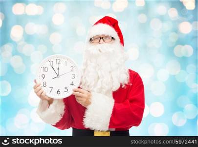 christmas, holidays, time and people concept - man in costume of santa claus with clock showing twelve over blue holidays lights background