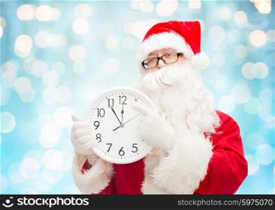 christmas, holidays, time and people concept - man in costume of santa claus with clock showing twelve pointing finger over blue holidays lights background