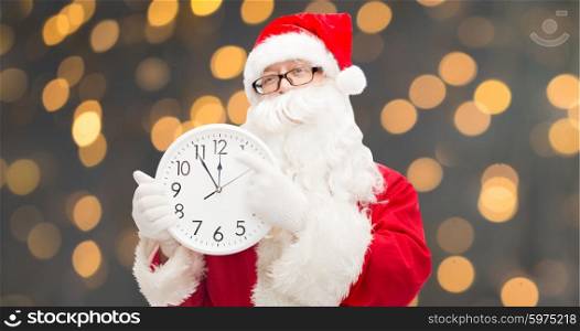 christmas, holidays, time and people concept - man in costume of santa claus with clock showing twelve pointing finger over golden lights background