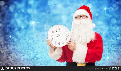 christmas, holidays, time and people concept - man in costume of santa claus with clock showing twelve over blue glitter or lights background