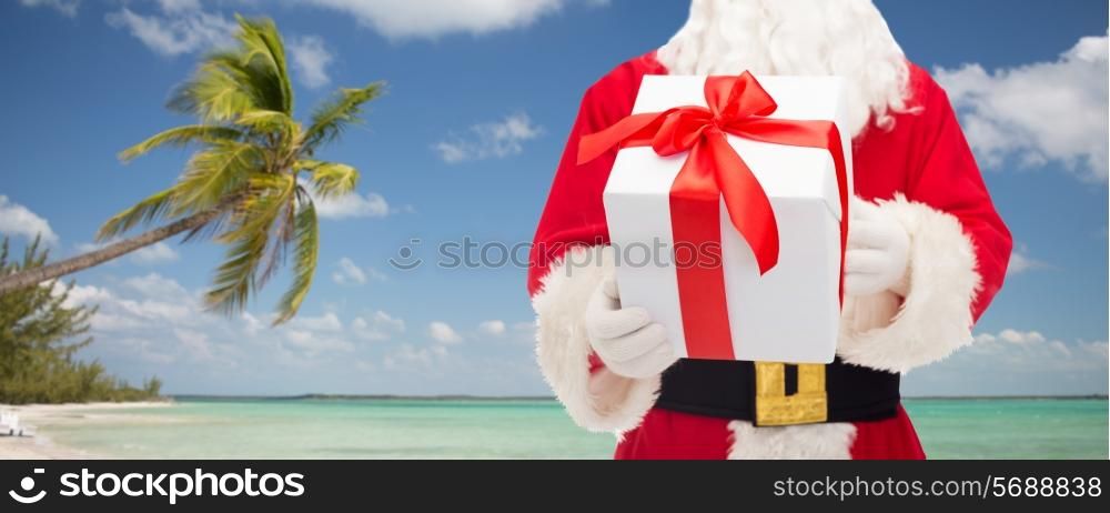 christmas, holidays, travel and people concept - close up of santa claus with gift box over tropical beach background