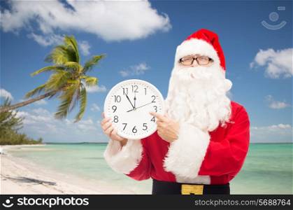 christmas, holidays, travel and people concept - man in costume of santa claus with clock showing twelve pointing finger over tropical beach background